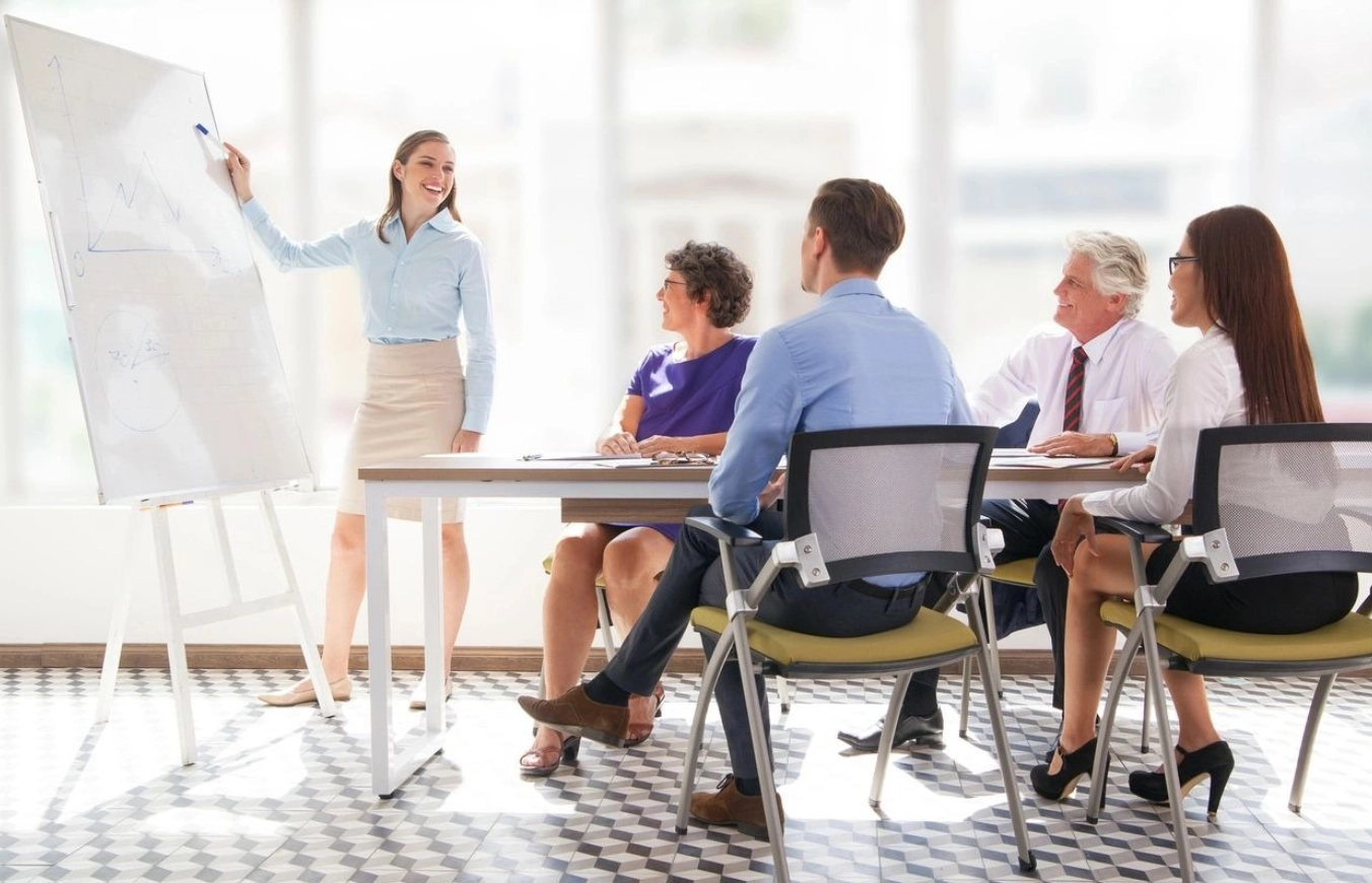 A woman standing at the front of a room with other people.